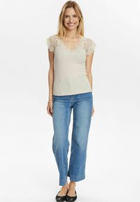 Top fitted color beige chiaro con maniche a cappuccio in pizzo, abbinato a jeans a gamba larga blu medio e scarpe flat nere. Texture liscia, design casual.