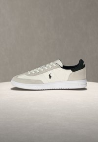 Pantof sport alb și gri deschis, cu talpă joasă, căptușeală interioară neagră, șireturi și logo cu calul Polo Ralph Lauren pe lateral și în zona călcâiului.