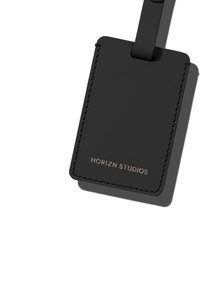 Étiquette de bagage en cuir noir avec une texture lisse, des bords arrondis et une sangle avec un bouton en métal. "HORIZN STUDIOS" embossé en argent.