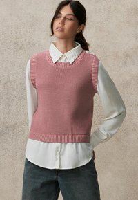Pull en tricot sans manches de couleur rose clair avec un motif texturé ; porté par-dessus une chemise blanche à col, associé à un jean en denim foncé.