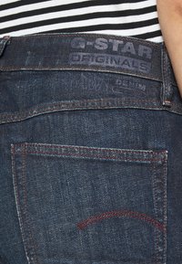 Tmavě modré džíny z denimu s logem G-Star Originals na zadním pasu. Mají kontrastní červené švy a klasický design kapes.