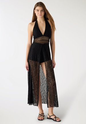 Młoda kobieta w czarnej koronkowej sukience na szyję bez rękawów z głębokim dekoltem w kształcie litery V i przednim rozcięciem, w czarnych sandałach na białym tle.