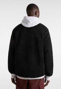 Chaqueta de forro polar negra con un exterior texturizado y suave, presentada con una sudadera gris debajo. El ajuste es relajado, con un cuello redondo y hombros caídos.