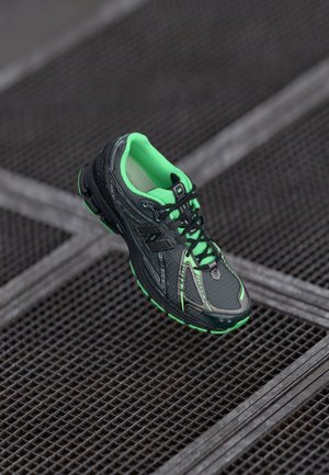 Schwarzer und neongrüner New Balance Laufschuh, der über einem Metallrost mit Kreuzmuster schwebt.