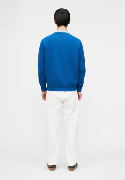 Άνδρας που στέκεται με την πλάτη γυρισμένη, φοράει μπλε πουκάμισο με μακριά μανίκια, λευκό παντελόνι και σκουρό πράσινα παπούτσια σε απλό λευκό φόντο.