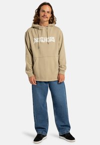 Beige hoodie with a front pocket, featuring white logo text. Paired with loose blue denim jeans and black sneakers.