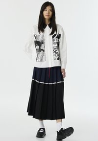 Camicia bianca con colletto nero e stampa grafica, abbinata a una gonna plissettata blu navy e nera con bordo in pizzo. Scarpe da ginnastica nere indossate.