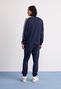 Navy tracksuit made of smooth fabric, featuring a light gray stripe along the shoulders. Pants have elastic cuffs, paired with white sneakers.