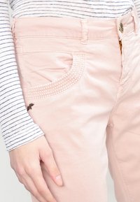 Jasnoróżowe spodnie z ozdobnym wzorem kieszeni z haftowanymi detalami. Materiał wydaje się miękki i ma gładką teksturę.