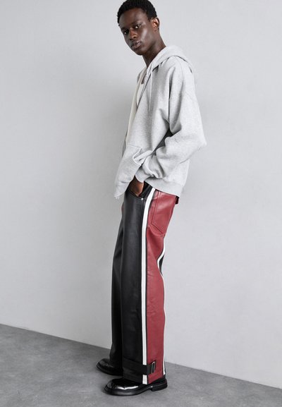 Un sweat à capuche gris avec une poche frontale, assorti à un pantalon en cuir à jambes larges rouge et noir, avec des rayures blanches sur les côtés et des accents en métal.