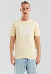 Jonge man in een lichtgele t-shirt met wit palmbomenontwerp en lichtblauwe ripped jeans, staande tegen een effen grijze achtergrond.