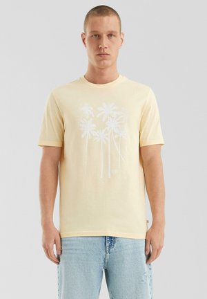 Jonge man in een lichtgele t-shirt met wit palmbomenontwerp en lichtblauwe ripped jeans, staande tegen een effen grijze achtergrond.