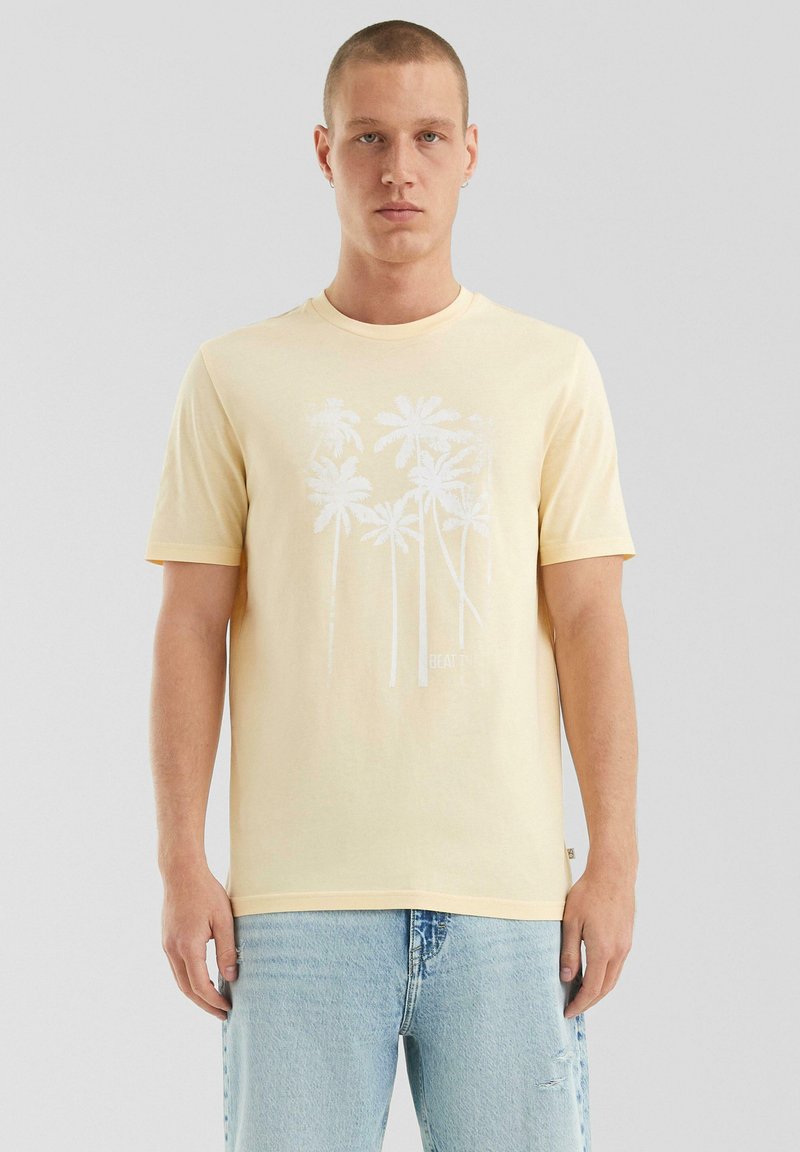 Jonge man in een lichtgele t-shirt met wit palmbomenontwerp en lichtblauwe ripped jeans, staande tegen een effen grijze achtergrond.