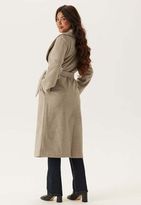 Beige Wollmischung langer Mantel mit einem Gürtel, aufgesetztem Kragen und seitlichen Taschen, kombiniert mit dunklen, ausgestellten Jeans und schwarzenPlateauabsätzen.