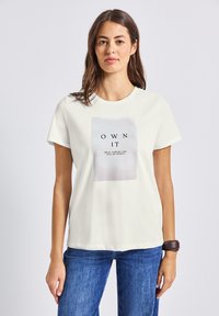 Femme portant un T-shirt blanc avec le texte "OWN IT" et un jean bleu, se tenant devant un fond clair, avec un bracelet noir au poignet gauche.