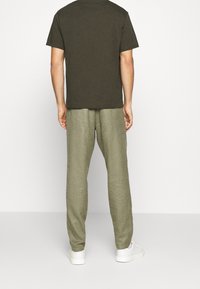 Miestenská móda s hnedým tričkom s krátkym rukávom a svetlozelenými ľanovými nohavicami, doplnenými bielymi teniskami, zobrazená zozadu.