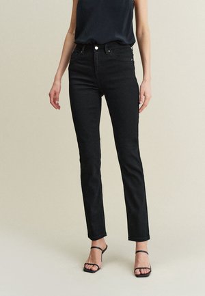 Femme portant un jean slim noir taille haute, un top noir sans manches et des sandales à talons noires à lanières, debout devant un fond uni.