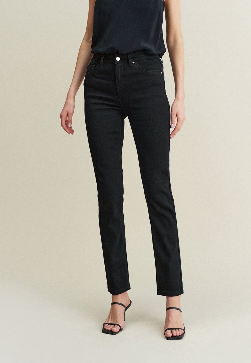 Femme portant un jean slim noir taille haute, un top noir sans manches et des sandales à talons noires à lanières, debout devant un fond uni.