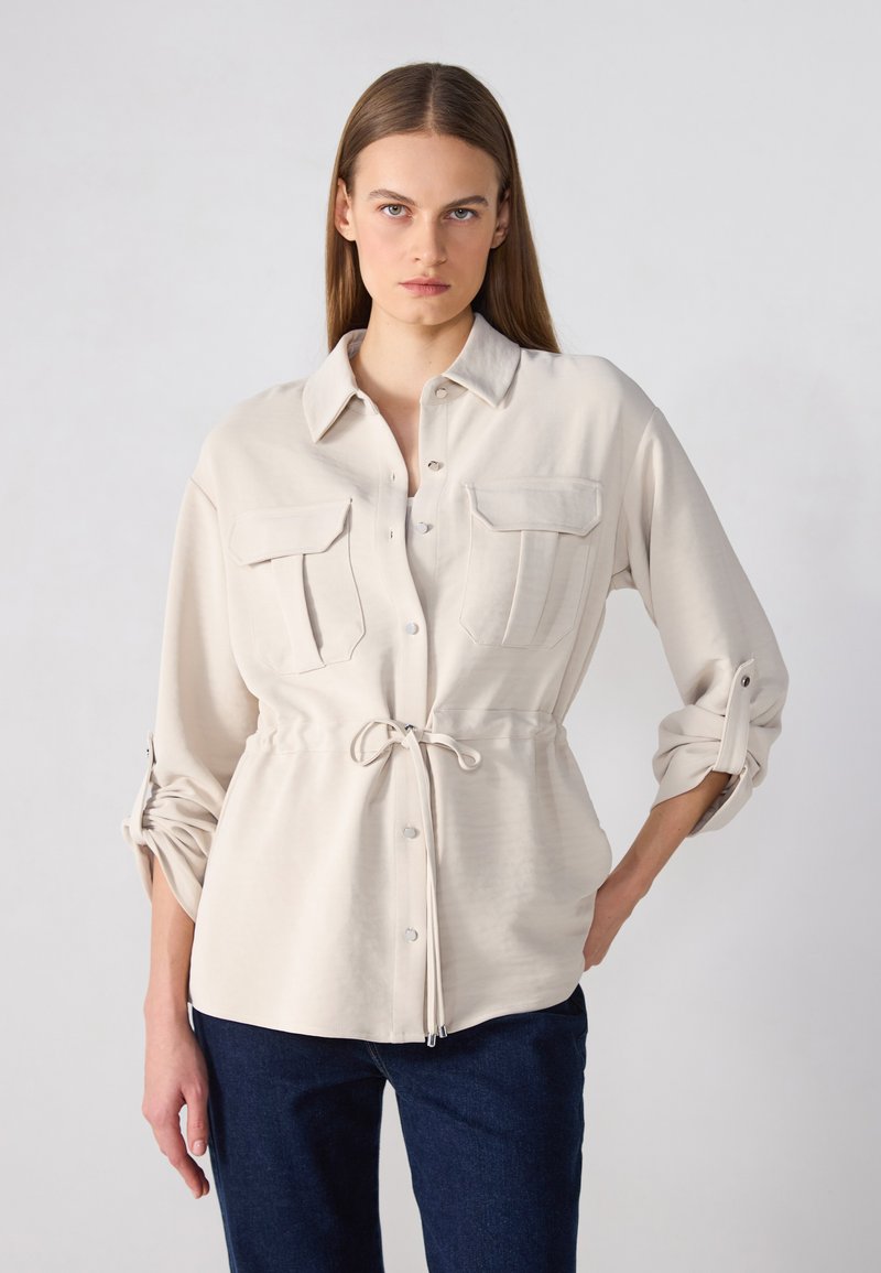 Lichtbeige shirt met een kraag, twee borstzakken en een rijgsluiting in de taille. Lange mouwen zijn opgerold. Gladde textuur en knoopsluiting.