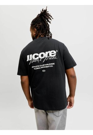Mand med dreadlocks iført en sort T-shirt med hvid tekst på ryggen og lysegrå jeans, der står foran en ensfarvet lysegrå baggrund.