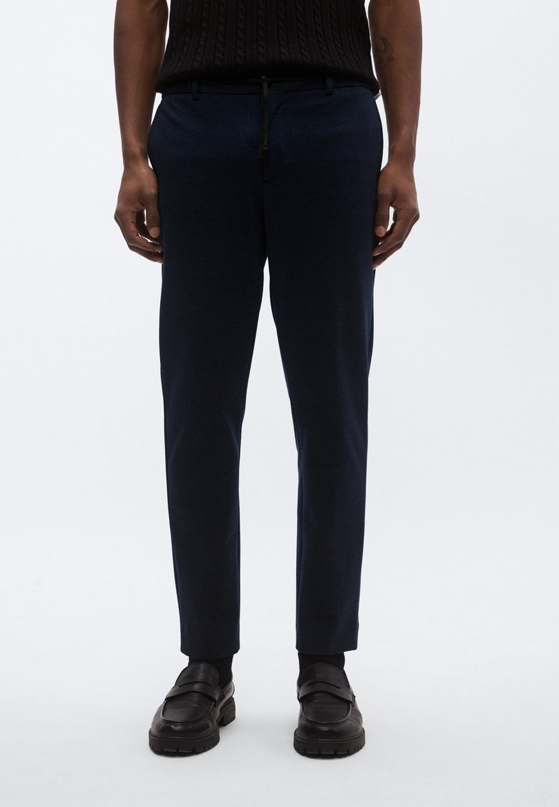 Παντελόνια σε σκούρο μπλε ναυτικό χρώμα με λεία υφή, διαθέτουν κορδόνι στη μέση και στενή εφαρμογή, συνδυασμένα με μαύρα loafers.