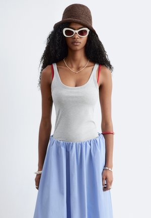 Femme aux longs cheveux bouclés portant un chapeau bob marron en tricot, des lunettes de soleil blanches, un débardeur gris, une jupe bleue et des bretelles de soutien-gorge rouges superposées.