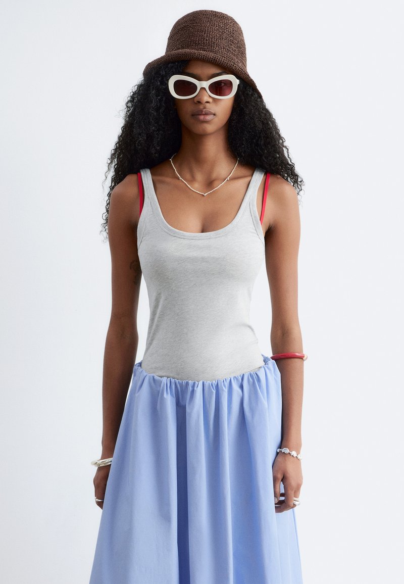 Femme aux longs cheveux bouclés portant un chapeau bob marron en tricot, des lunettes de soleil blanches, un débardeur gris, une jupe bleue et des bretelles de soutien-gorge rouges superposées.