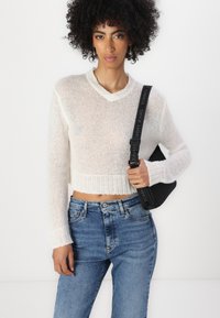 White, cropped knit sweater with a V-neck and ribbed hem, paired with light blue high-waisted jeans and a black bag with a branded strap.