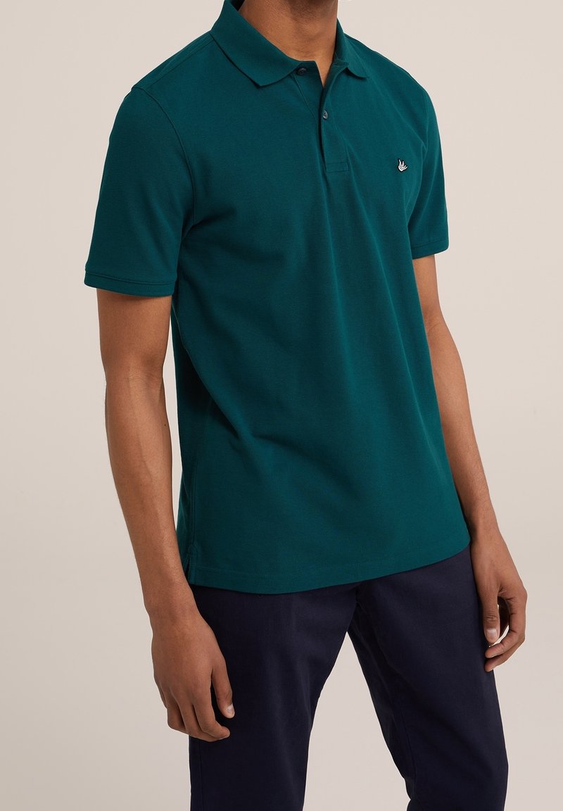 Polo bleu sarcelle en coton avec manches courtes, un col et un petit logo sur la poitrine. Associé à un pantalon foncé.