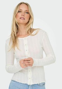 Witte geribde cardigan met een knoopsluiting aan de voorkant, golfkanten manchetten en een subtiele streepjespatroon. De stof lijkt lichtgewicht en gestructureerd.