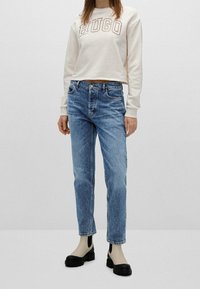 Sudadera crema con el logo "HUGO", combinada con jeans de mezclilla azul, de cintura alta, pierna recta y zapatos de plataforma negros con detalles recortados.