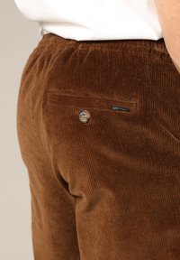 Pantalon en velours côtelé marron avec une finition texturée, doté d'une poche arrière et d'un bouton en métal. Comprend une étiquette de marque visible.