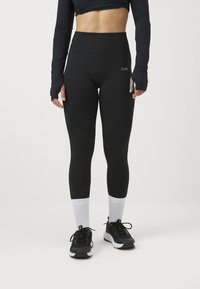 Leggings noirs taille haute avec une texture lisse et un subtil détail en relief, associés à des chaussures de sport noires et des chaussettes rayées grises.