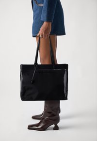 Black tote bag with a smooth texture, featuring leather handles, a logo, and a mesh panel on the side, held by a person wearing a blue jacket.