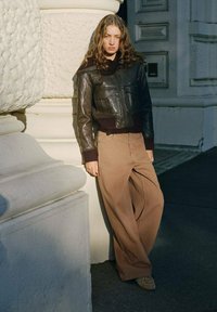 Zwarte leren cropped jack met voorkruiken, gecombineerd met lichtbruin wijde broek. Achtergrond tegen een gestructureerde witte kolom.