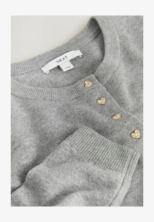 Pelēks adīts kardigans ar ribotu apkakli un piedurknēm, kuram ir trīs zelta sirds formas pogas priekšējā daļā. Redzama zīmola birka.