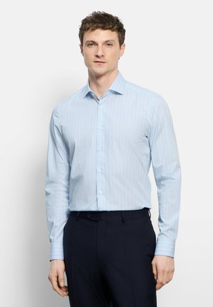 Homme portant une chemise à rayures bleu clair à manches longues, rentrée dans un pantalon formel sombre, sur un fond uni.