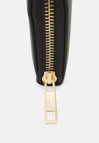 Schwarze Reißverschlusstasche mit einem goldenen ZIP-Zipper, die eine strukturierte Oberfläche und ein schlankes Design aufweist. Die Tasche bietet eine elegante, funktionale Aufbewahrung.