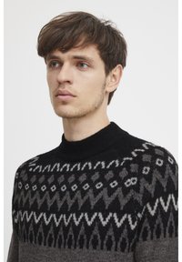 Strickpullover mit einem schwarz-, grau- und weiß-geometrischen Muster. Design mit hohem Kragen, strukturiertem Stoff und verschiedenen Farbtönen, die einen Kontrast erzeugen.