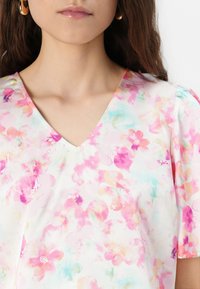 Blusa con estampado floral que presenta un escote en V, mangas cortas abullonadas y colores pastel con flores en rosa, melocotón y azul claro sobre un fondo blanco.