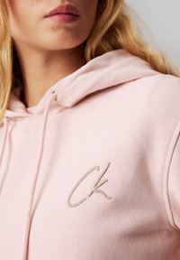 Lys rosa hettegenser laget av myk stoff, med en fremtredende beige brodert "CK"-logo på forsiden.