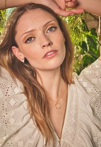 Zelta kaklarota ar apaļu akcentu, nēsāta virs baltas, teksturētas blūzes. Garas matu, vasaras raibumi uz ādas un maiga apgaismojuma piešķir vizuālu pievilcību.