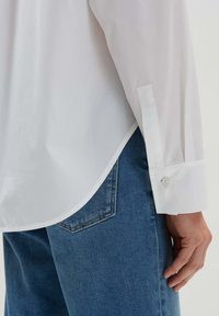 Camicia bianca con maniche lunghe, tessuto liscio, polsini con bottone, orlo curvo, abbinata a jeans azzurri chiari con tasca posteriore e cuciture sottili.