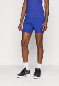 Pantalones cortos deportivos azules con forro incorporado, cinturilla elástica y costuras en contraste. Combinados con una camiseta sin mangas azul a juego y zapatillas deportivas negras.