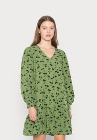 Vestido verde com decote em V, mangas longas abalonadas e bainha em camadas. Apresenta um padrão de cerejas pretas por todo o lado. Tecido de textura suave.