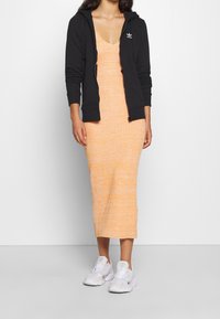 Svart zip-up huvtröja lager över en texturerad orange midi-klänning, bärs med vita sportskor. Klänningen har en figursydd silhuett.