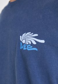 Tamsiai mėlynas medvilninis marškinėlis, su baltu ir mėlynu grafiniu dizainu, vaizduojančiu bangos formos raštą, ir žodžiu "LEE" kairiajame krūtinės šone.