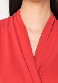 Punainen kietaisupaita, joka on tehty sileästä kankaasta ja jossa on syvä V-aukko sekä päällekkäiset etukappaleet, jotka korostavat kankaan tekstuuria.