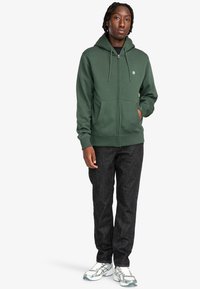 Cazadora con cremallera de color verde oscuro, con cordones, bolsillos frontales y puños acanalados, combinada con jeans negros y zapatillas deportivas blancas.