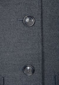 Testura di cappotto in lana grigia caratterizzata da due grandi bottoni lucidi con quattro fori ciascuno. La cucitura è visibile lungo il posizionamento dei bottoni.